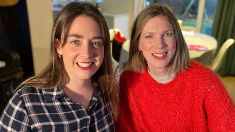 Two ladies smiling at the camera. On the left Gemma Holbird wearing a chequered buttoned shirt. She has highlighted long brown hair. On the right is Sarah Jane Sauntson with shoulder-length straight blonde hair wearing a bright red oversized woollen jumper.