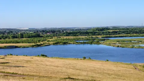 RSPB RSPB St Aidan's reserve near Leeds with a blue lake surrounded by yellow and green grassy fields with bushes and reeds.