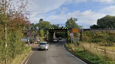 Streetview image of a short stretch of single carriageway road in a rural area, running under a heavily sign posted railway bridge.