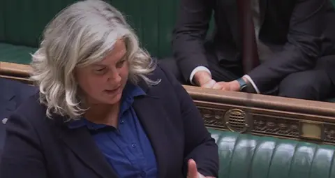 A woman wearing a blue shirt and a black blazer. She is standing in the House of Commons and behind her are dark green benches