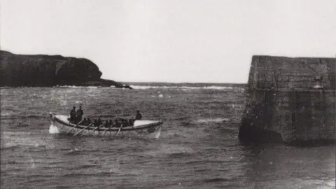 RNLI An old black and white picture showing a wooden longboat, there are 5 oars on the boat. they are just at the edge of the harbour.
