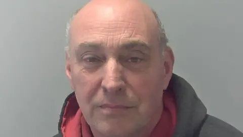 Suffolk Police Matthew Bye's police custody image. He stares at the camera. He is largely bald with some grey hair on the sides of his head. He wears a grey hoodie that has a red lining.