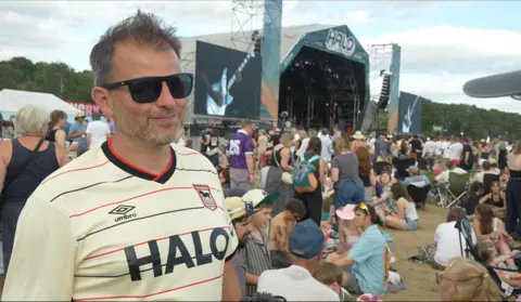Connor Bennett/ BBC Paul Hamilton smiles, wearing an Ipswich Town Jersey, which has Halo emblazoned as the club sponsor across the chest. Festivalgoers and the main stage are the background.