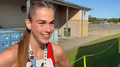 MANX RADIO Aimee Christian smiling. She has long light brown hair, which is tied back, and is wearing a red team Isle of Man vest with a number on it. She is standing on grass and there is a brick-built building behind her.
