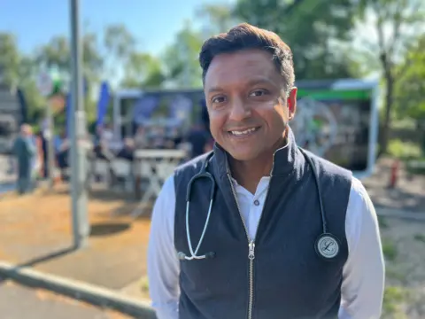 Navin Chandra, a consultant cardiologist, stands in front of a pop-up clinic in Slough. He is wearing a white shirt with a grey sleeveless jacket. He has a stethoscope over his shoulders and is smiling.  