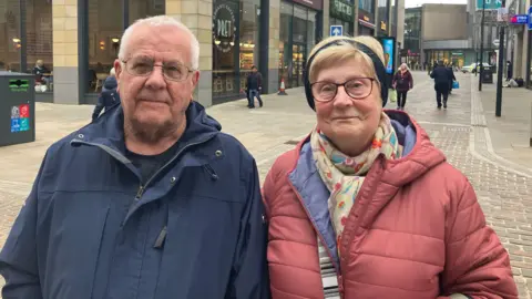 BBC/Grace Wood A man wearing a blue rain jacket with glasses and short grey hair and a woman in a pink puffer coat with a headband, blonde hair and glasses.
