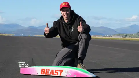 The image shows A man with a red baseball cap and a black hoodie, giving a double thumbs-up, kneeling behind a remote control car on a long stretch of road.