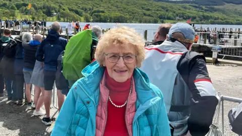 Anne Hall looking into the camera as she stands on the shore of Coniston Water. She is wearing glasses and a light blue coat, and has fair hair.