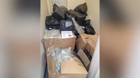 A small room with full black bin bags and brown cardboard boxes piled on top of each other. 
