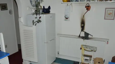 Cambridgeshire Police A kitchen which has small doors to the hall, and contains a fridge/freezer. There is a radiator with a charred tea towel on top and charring on the wall above