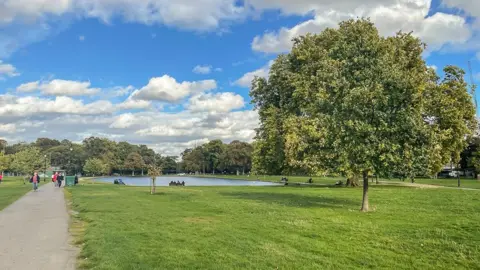 File photo of Clapham Common in the daytime. There is green grass, some trees and people walking on a path and sitting on a bench looking at a pond.