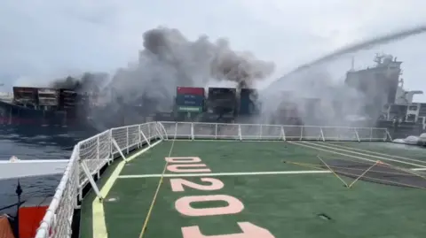 A ship is on fire. Thick dark smoke rises from the containers on its deck. In the foreground, we see the deck of another vessel with railings and a large green area marked “0205.” A strong stream of water sprays from the boat, aiming at the fire. 