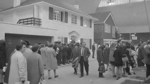 Crowd gathered around a model two-storey house inside a large exhibition hall in a black and white image.