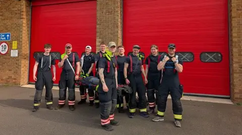 Cambridgeshire Fire and Rescue Nine young men and women wearing dark coloured t-shirts and trousers with fluorescent stripes towards the bottom standing in a group with a stretcher carrying a dummy wrapped in dark clothing. There are large red doors behind them on a red brick building. 