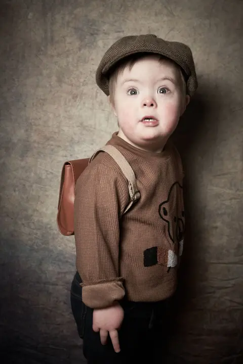 Debbie Todd Albie, a young boy, wears a brown flat camp and brown jumper with a teddy bear design on it with a satchel on his back. 