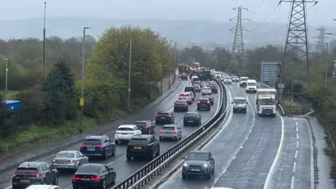 A traffic jam with cars backed up along the road. Tractors can be seen in the distance. One side of the road heading one way is full of cars; the other side heading the other way is a bit clearer.
