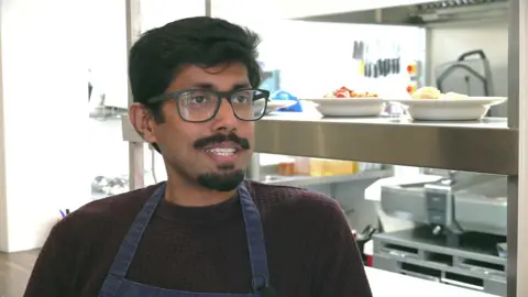 A man in a dark jumper and blue apron looking to the right of the frame. Silver kitchen equipment and a couple of plates of food are in the background.