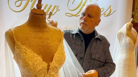 Huw Rees Photo of Huw Rees in his store window display, you can read the gold signage in the window reading "Huw Rees". Huw is holding a veil which is attached to a mannequin dressed in a strappy lace white wedding dress. He looks with concentration at the shoulder strap. He has silver hair and a silver beard. He wears a black turtleneck top on with a black and white houndstooth shacket. Another mannequin can be seen in the background, with an embellished dress and white veil. 