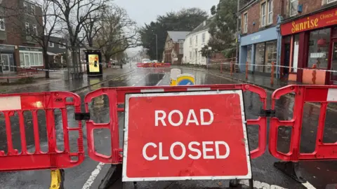 Red road closed sign in Exeter. Fallen tree leaning on a house can be seen in the distance