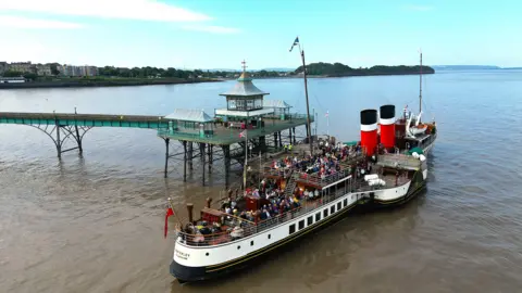 Waverley Excursions A large old-fashioned paddle steamer is moored at the end of a pretty green pier in a wide channel of water.