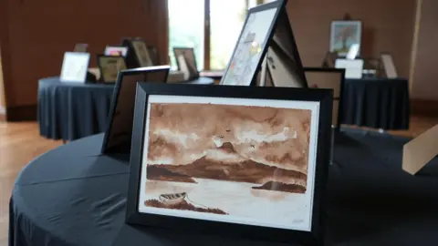 A watercolour painting of an island with a hill and a moody sky in colours of sepia and brown in a black frame. It is on a table with a black cloth with other paintings behind it and on other tables in the background. It is a large room with a window in the background.