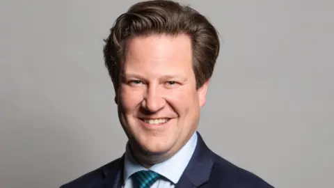 LDRS A man in a navy blue suit and a pale blue shirt with a green tie, smiling against a grey backdrop.