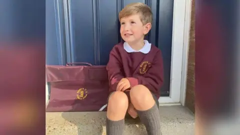 Family handout A young boy with dark blond hair is sitting on a step outside a blue door. He is wearing a burgundy school uniform with long grey socks and black shoes. The school uniform's jumper has a yellow circular logo, and he has a matching school bag next to him. He is looking up to the side and smiling.