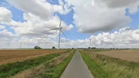 A Google street view image of a narrow, grey, country lane with a ditch at either side of it. The road is surrounded by large farmland fields and several wind turbines are visible in the distance.