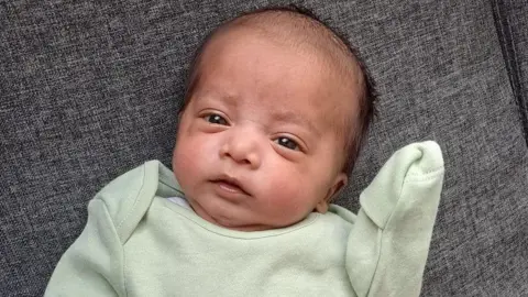 A young baby looks at the camera. He has dark brown hair and wears a light green baby grow.