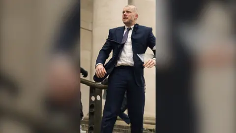 Actor Russell Tovey as Barclay in a navy blue suit. His tie is being swept by the wind and he is looking into the distance