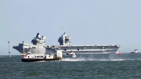  ALI TRE HMS Prince of Wales is a light grey aircraft carrier - her flight deck is 70 metres wide and 280 metres long – enough space for three football pitches. She can be seen at sea with several small boats clustered around it