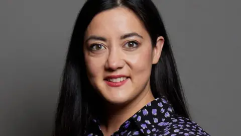 UK Parliament Sarah Owen MP looks directly at the camera. She has long dark hair, red lipstick and is wearing a black and mauve patterned blouse. Behind her is a plain grey backdrop.