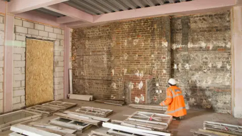 UCL Archaeology South-East A man in orange high-vis clothing and a hard hat examines an old-looking brick wall with a bricked up fireplace. The other walls and roof appear to be newer. Construction materials are seen on the floor of the room.