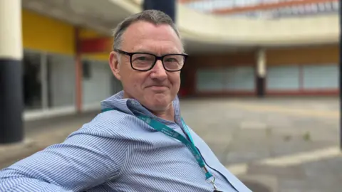 Mike Stonard is sitting on a bench in Anglia Square. He's wearing a blue and white pinstripe shirt along with a light blue lanyard. He also is wearing glasses.