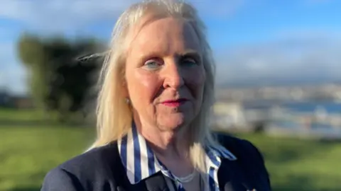 Julie Edge, a woman with light blonde hair, she wears a blue and white stripy shirt and looks seriously at the camera.