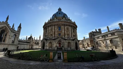 BBC/Curtis Lancaster Historic circular stone building with a large domed roof at the center of a university courtyard. The domed structure is surrounded by Gothic and medieval-style academic buildings with arched windows, towers, and decorative stonework. A black metal fence encloses a grassy lawn around the central building, while several bicycles are parked along the fence and nearby cobblestone paths. The scene is brightly lit under a clear blue sky, with the sun positioned behind the dome, creating a subtle halo effect around the roof.