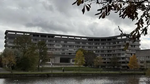Steve Hubbard/BBC A large, curved concrete building with multiple rows of windows stands behind a grassy area and a pond. The building is Bedford Borough Council's headquarters. Several trees with autumn foliage are scattered across the scene, and branches frame the top of the image. The sky is overcast, creating a moody atmosphere, and a few people can be seen walking along the path near the water.