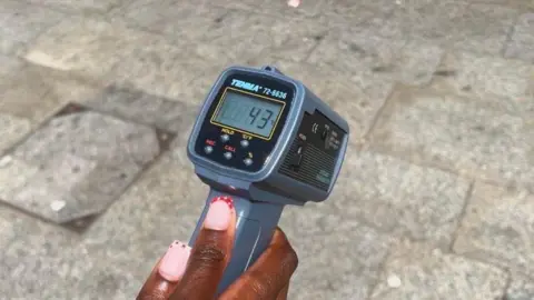 Hand holding a handheld infrared temperature gun over a paved stone surface outdoors. The digital display on the device shows a temperature reading of approximately 43°C, indicating the heat of the pavement. The ground is made of large grey stone slabs, with a utility cover visible in the background.