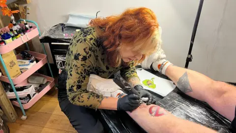 Tattoo artist Holly Dosdale at work. She is working on a tattoo on a man's leg. 