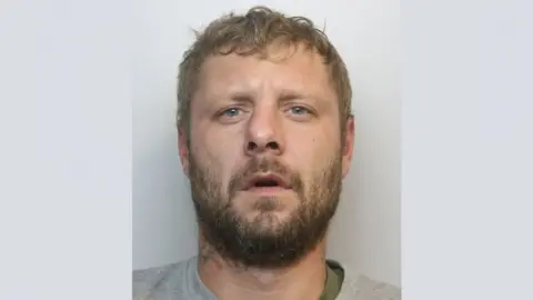 A police mugshot of Joshua Whittaker. He is wearing a green t-shirt underneath a grey crewneck sweatshirt. He has blue eyes, dirty blonde hair and a full beard. He is looking at the camera with his mouth slightly open.