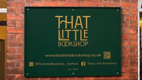A red brick wall of a book shop and a large forest green sign with gold writing. The writing reads "That Little Bookshop". it also has the shop's website and social media account handles on it, and at the bottom it says "established 2024".