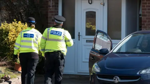 Two police officers, one male and one female, walk away from the camera towards the house of Sergei Skripal in Salisbury at the time of the Novichok poisonings. They are both wearing large yellow high-vis jackets. The house has a white front door and a dark blue Peugot car is parked outside