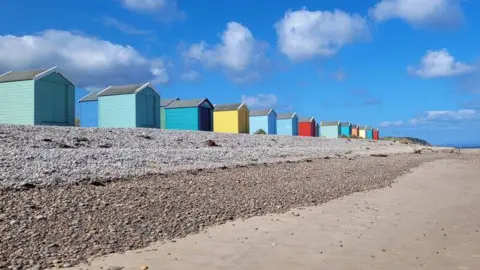 Nicky Thomas Colourful beach huts line up along a pebbly beach that becomes sandy closer to the water.