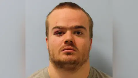 A mugshot of Jonty Bravery, who is wearing a grey shirt. He is staring at the camera and has a beard and short, blonde hair. 