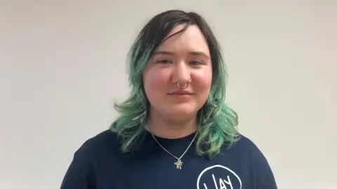 Girl with blue top and a cross necklace. She has a septum piercing and black hair with green coloured highlights. She smiles at the camera.