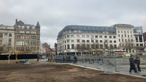 Fencing panels surround an area of Piccadilly Gardens in which soil can be seen.