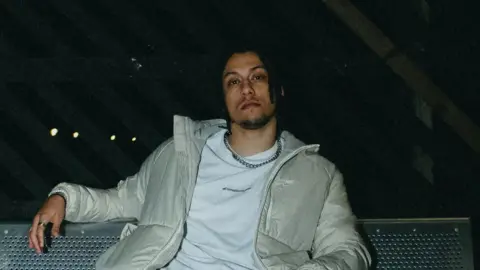 Max Jones sitting on a metal bench with a pattern created by small holes. he is wearing a cream jacket, a white top with a small black written logo and a silver chain. It is taken at night and in the background is a row of four lights 
