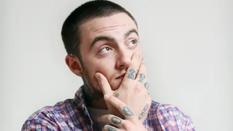 Getty Images Mac Miller pictured with his tattooed hand covering his face and looking up away from the camera. He wears a blue, white and pink plaid shirt over a white T-shirt and is photographed against a bare white wall. 