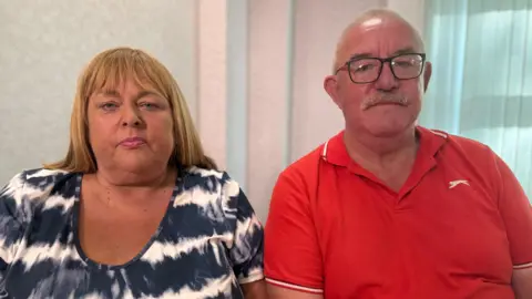 A woman, with long blonde hair, wearing a white and navy top. She is sitting beside a bald man who is wearing a red t-shirt and glasses.