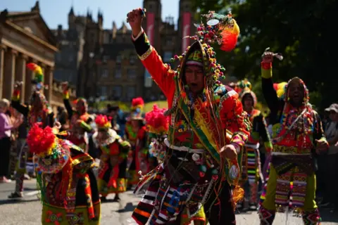 Parade participants wearing highly decorated multicoloured costumes with embroidered patterns, beads and feathered headdresses perform in a street procession. One performer is captured mid-dance in the foreground.